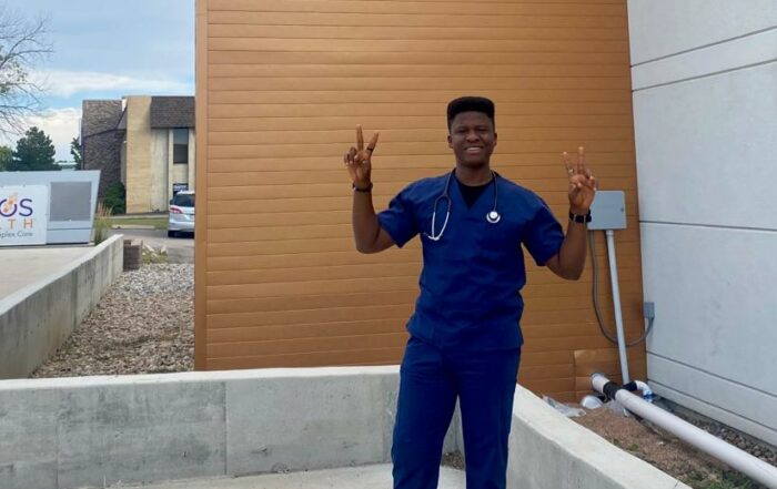 AMO Photo and Blog Winner from August 2023, Samuel, poses with a smile and two hands raised in front of a hospital building.
